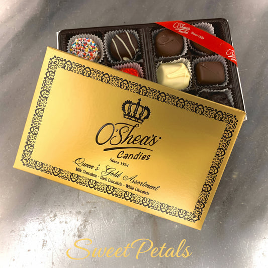 An open box of O'Shea's Candies 'Queen & Gold Assortment' milk chocolate, dark chocolate, and white chocolate truffles, with the gold and black ornate-patterned lid leaning in front of the black tray on a gray surface.
