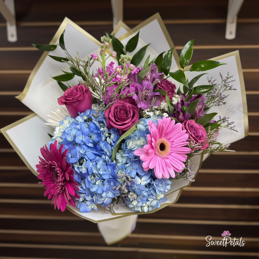 Close-up of deep purple roses blue hydrangea and pink gerbera daisies in white gold wrap – SweetPetals violet dream luxury bouquet